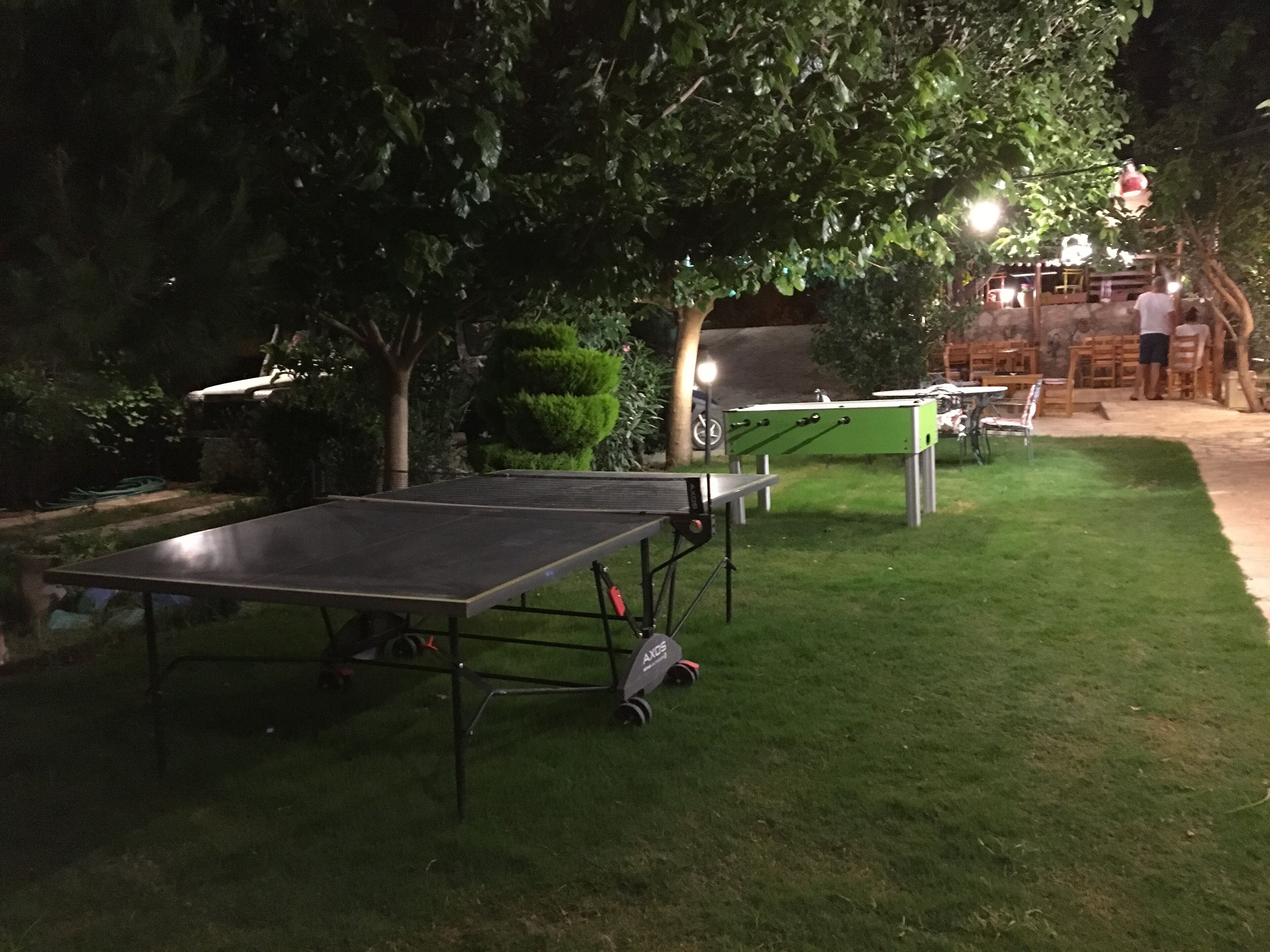 An outdoor area with a ping pong table and a foosball table. There are trees and a building with lights in the background.