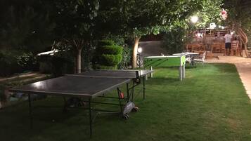 An outdoor area with a ping pong table and a foosball table. There are trees and a building with lights in the background.