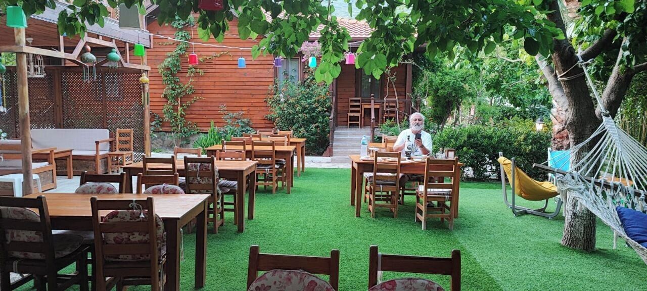 An outdoor dining area with wooden tables and chairs, surrounded by greenery and a hammock.