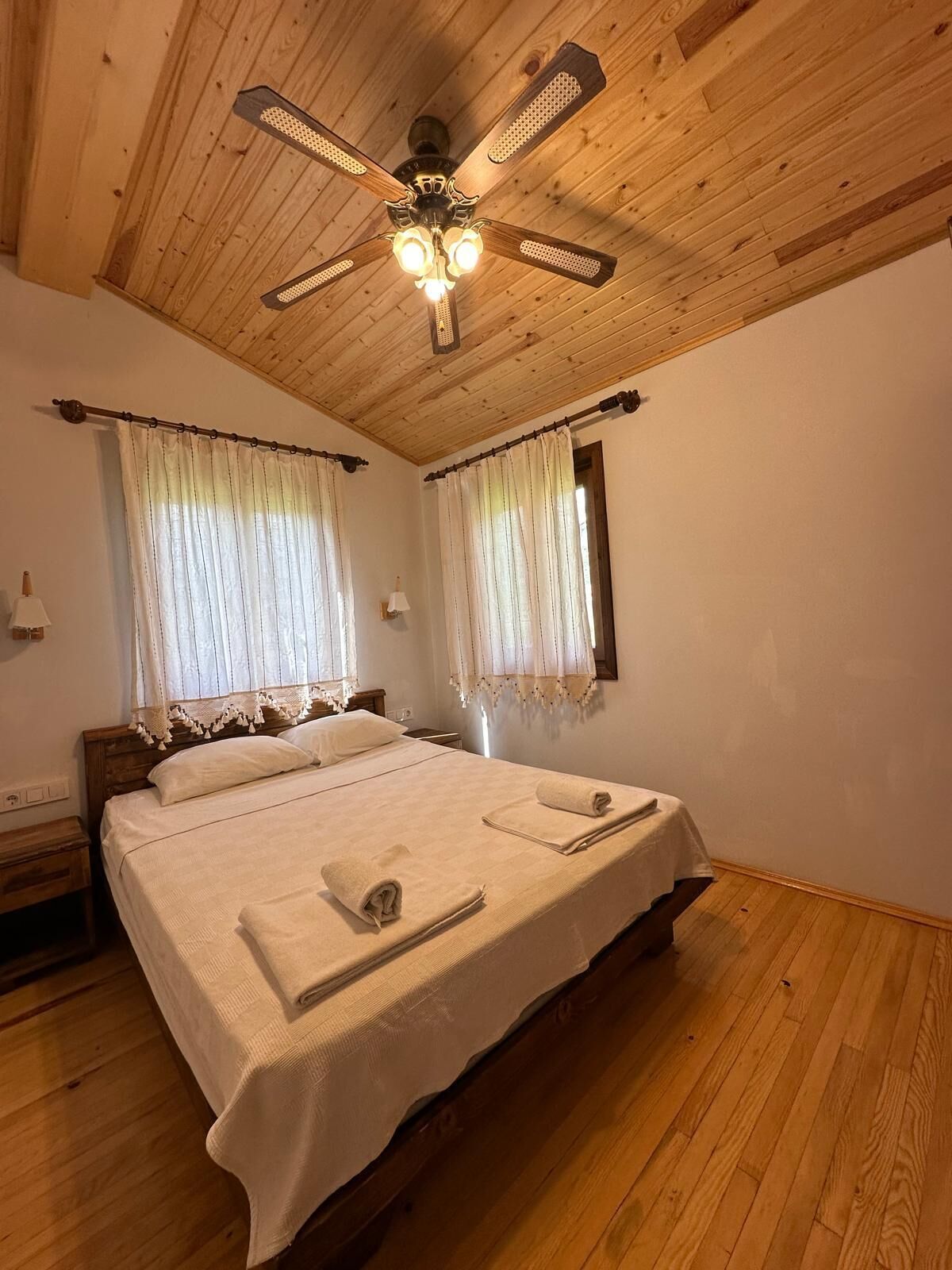 Un dormitorio con techo de madera, cama con ropa de cama blanca y dos ventanas con cortinas de gasa.