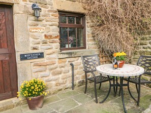 A stone cottage with a sign reading "MIDFEATHER COTTAGE," a round table with chairs, and potted yellow flowers.