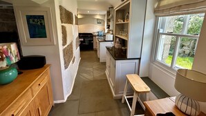 A narrow kitchen with white cabinets, a wooden bench, and a window with a view of greenery. - 14 Boscaswell Downs (Penzance)