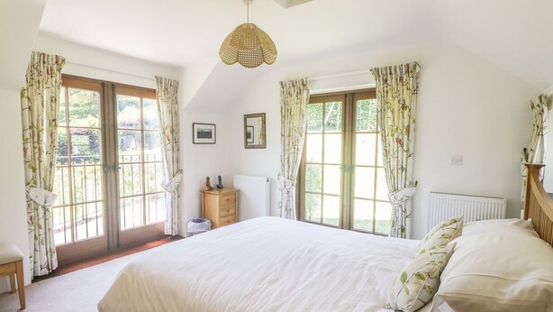 Un dormitorio con una cama grande, dos ventanas con cortinas, una mesita de noche de madera y una lámpara colgante.