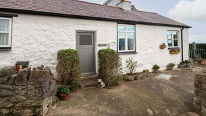 Une petite maison blanche avec une porte grise, une petite fenêtre et une cheminée. On y trouve des plantes en pot et un mur de pierre à l’avant.