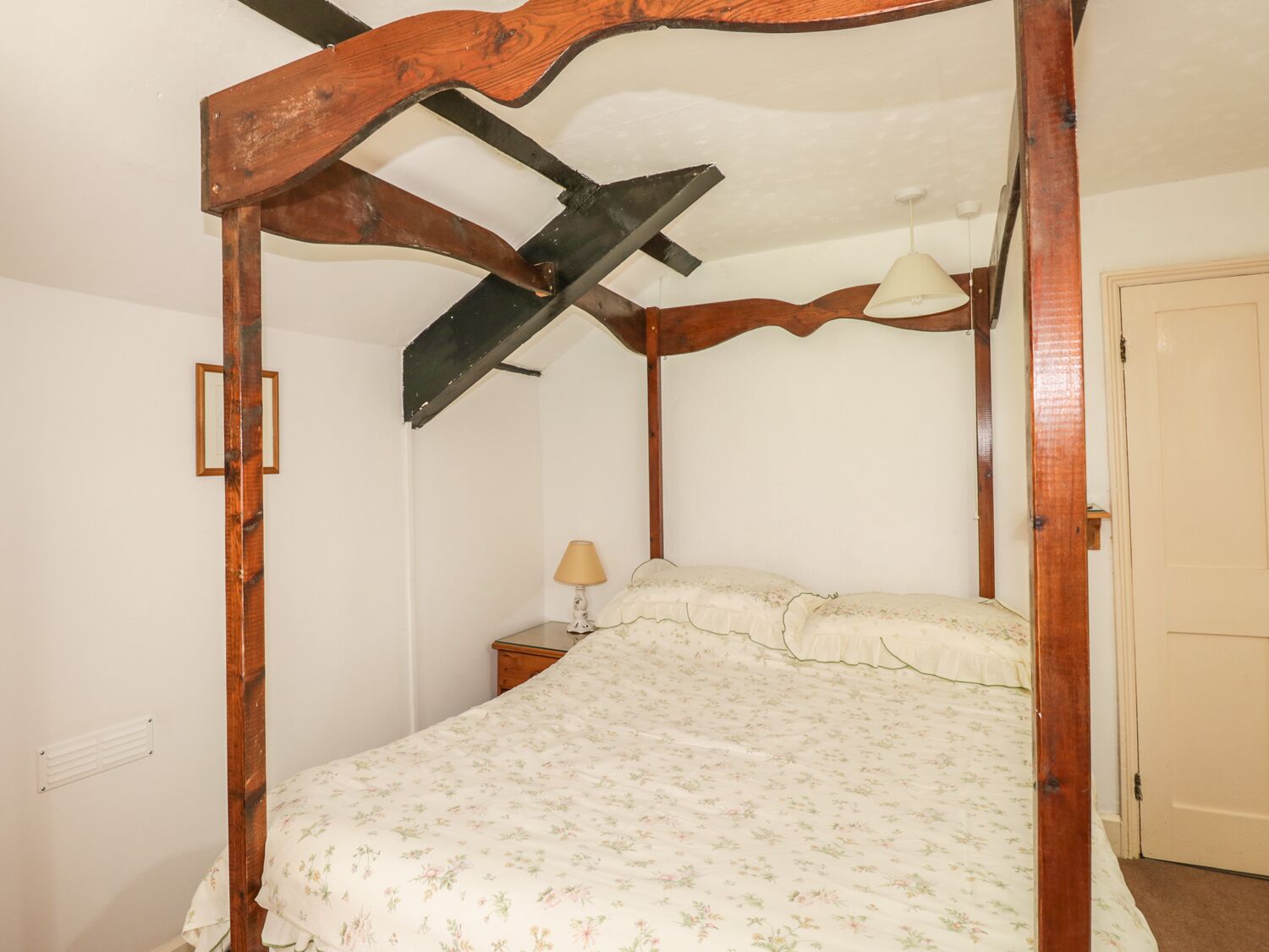 A four-poster bed with a floral bedspread, a nightstand with a lamp, and a door to the right.