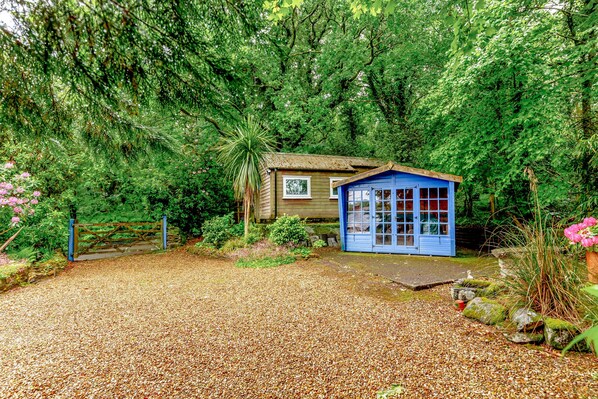 A rustic cabin with a gravel driveway, surrounded by dense greenery and a wooden fence.