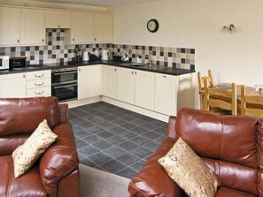 A compact kitchen with white cabinets, a black countertop, and a tiled backsplash. There is a dining table with chairs and a clock on the wall. - Ty Isaf Penrhyddion (Llanrwst)