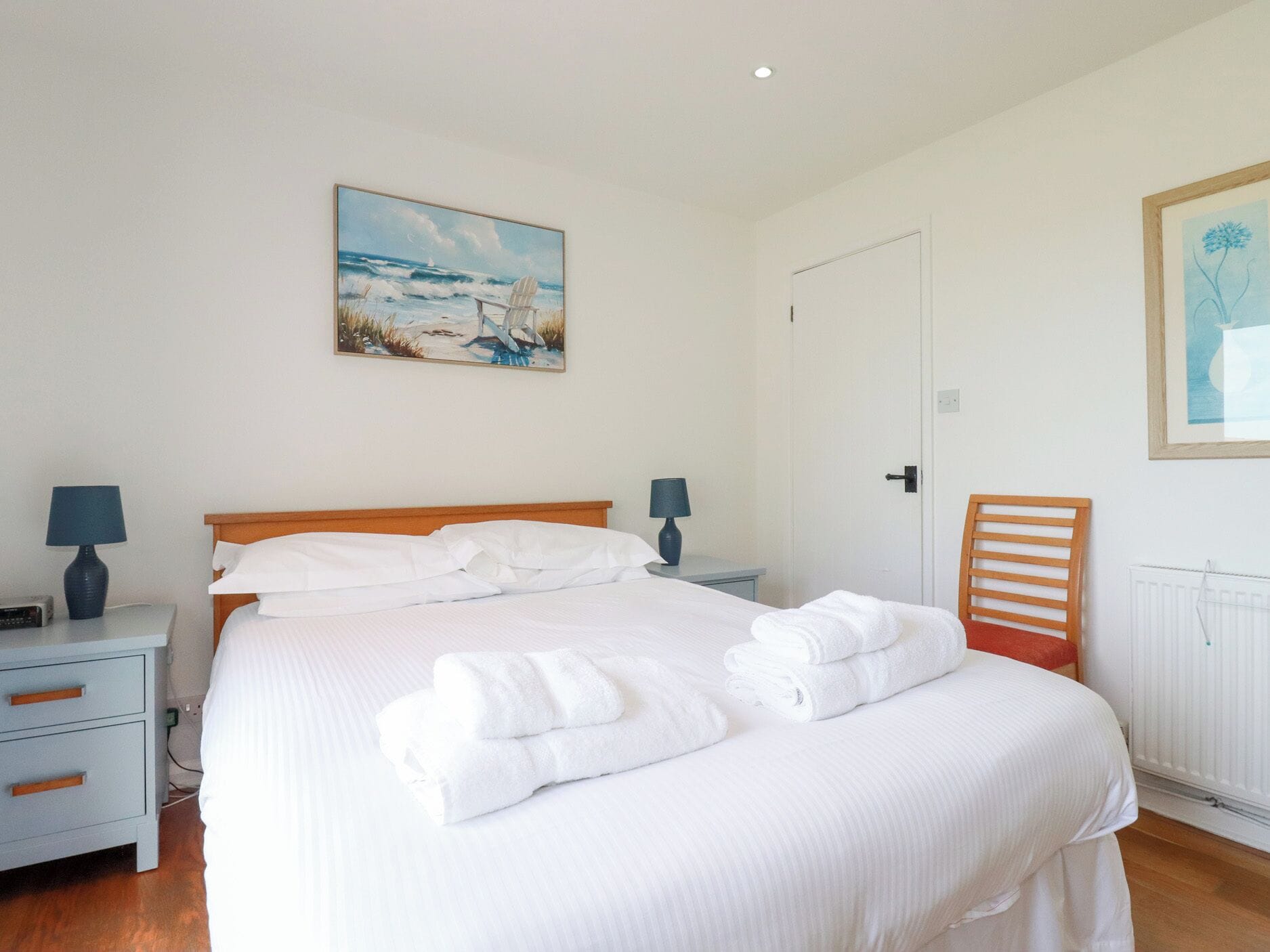 A neatly made bed with white linens and towels, a wooden headboard, a nightstand with a lamp, and a painting of a beach scene on the wall.