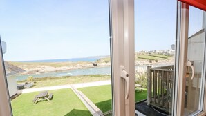 A view from a room with a large window overlooking a grassy yard with a wooden deck and a view of the sea and rocky coastline. - The Beach House (Newquay)