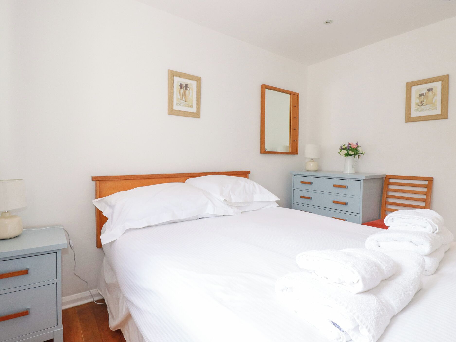 A neatly made bed with white linens, a wooden headboard, and a nightstand with a lamp. There are two framed pictures on the wall, a mirror, and a small table with a vase of flowers.