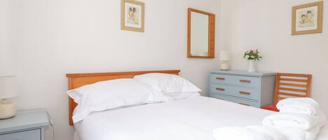 A neatly made bed with white linens, a wooden headboard, and a nightstand with a lamp. There are two framed pictures on the wall, a mirror, and a small table with a vase of flowers.