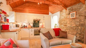 Una sala de estar acogedora con pared de piedra, techo de madera y una cocina con armarios blancos y un hervidor rojo.