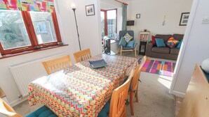 A dining area with a table covered by a patterned tablecloth, wooden chairs with cushions, a radiator, a window with curtains, a sofa with colorful cushions, and a bookshelf. - Kernow Cottage (Helston)
