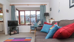 A living room with a sofa, colorful cushions, a TV on a stand, a chair, a window with curtains, and a view of houses outside. - Kernow Cottage (Helston)