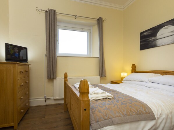 A bedroom with a wooden bed, a nightstand with a lamp, a dresser, a window with curtains, and a painting on the wall. - Roa Island House (Barrow-in-Furness)