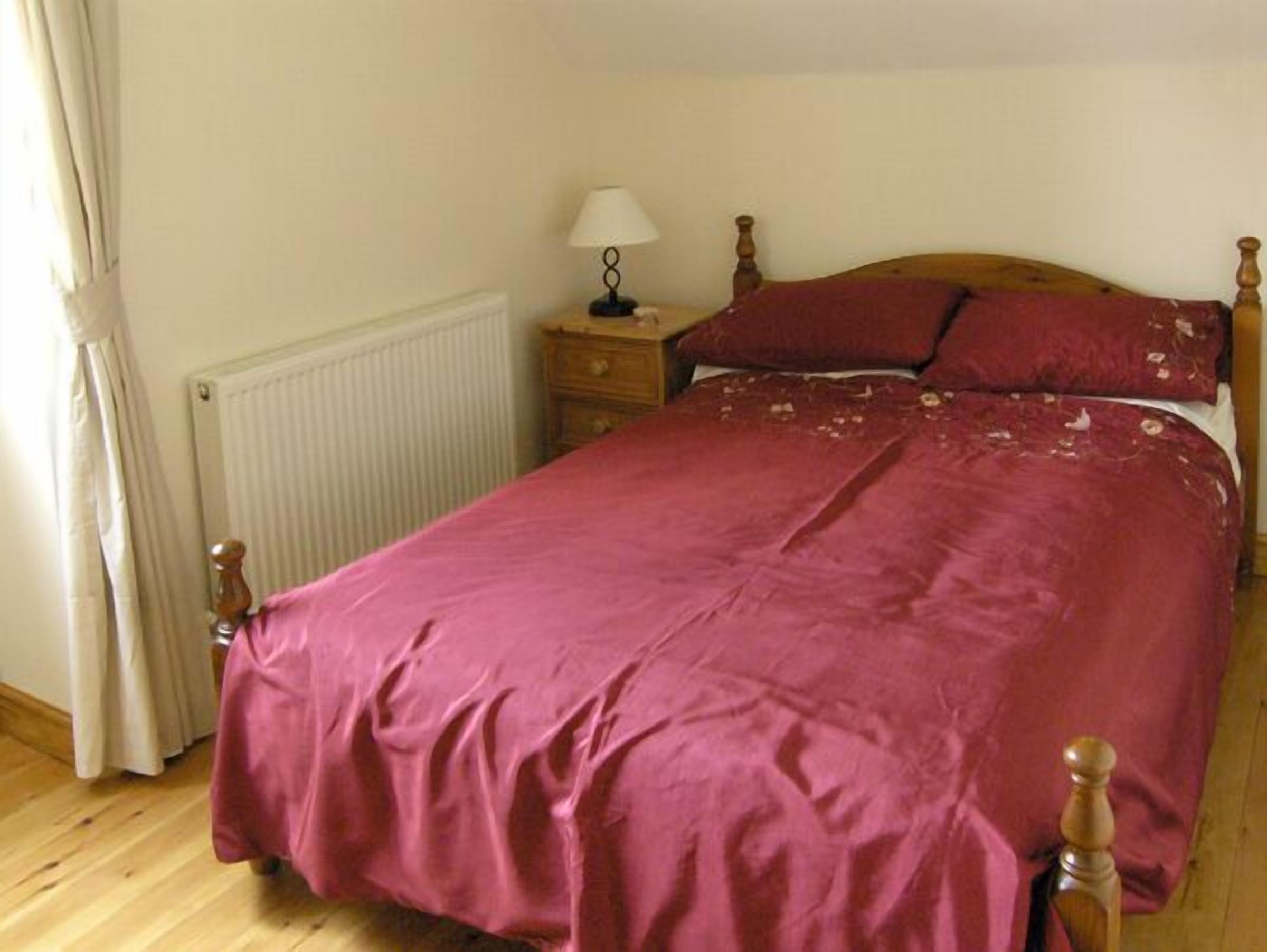 A bedroom with a wooden bed frame, a red bedspread, a bedside table with a lamp, and a radiator.