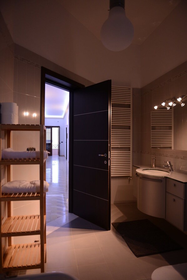 Un baño moderno con lavamanos, espejo y barra para toallas. Se ve un estante de madera con toallas, y una puerta conduce a otra habitación.