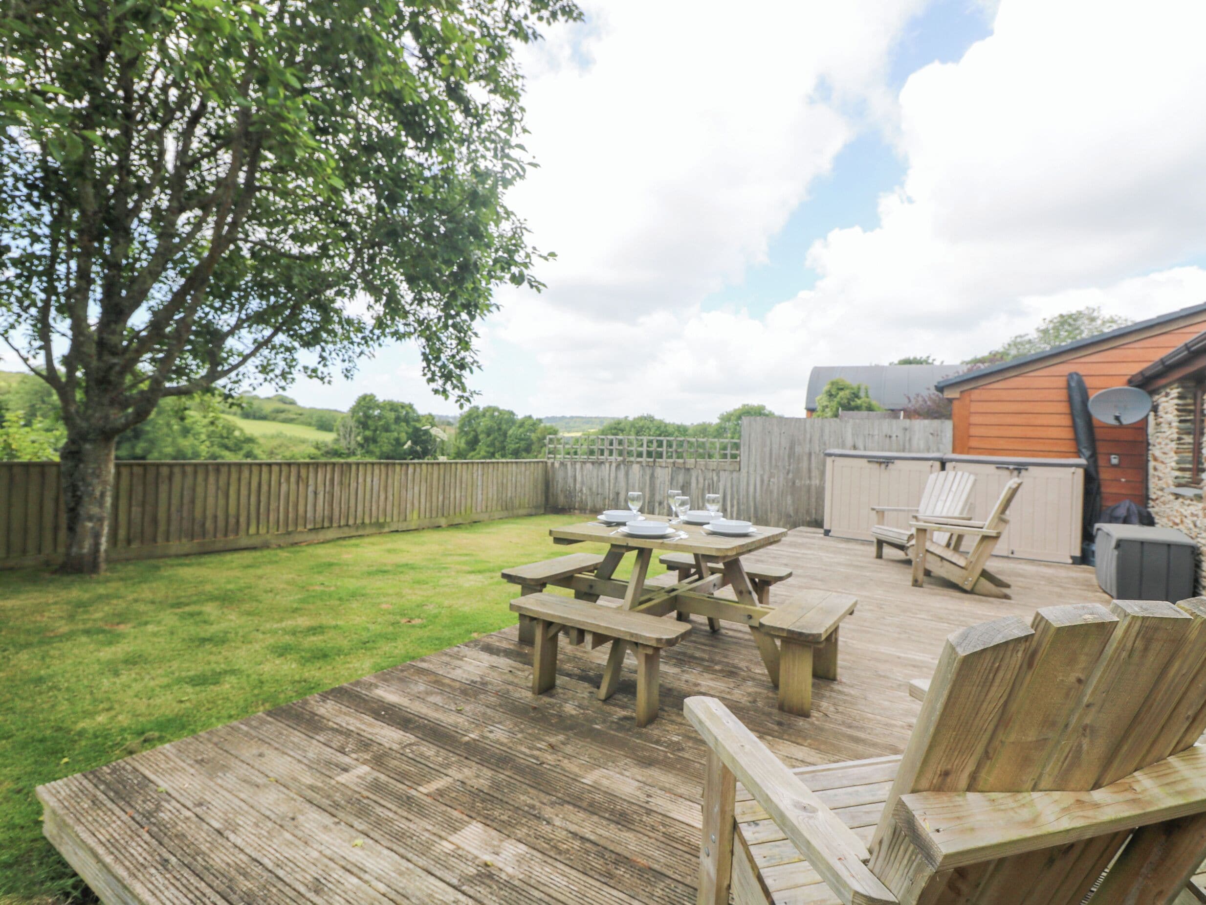 A wooden deck with picnic tables and chairs, surrounded by a wooden fence and a tree.