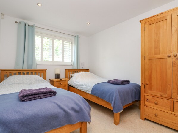 A bedroom with two wooden beds, a wardrobe, a window with curtains, and a bedside table with a lamp. - Rock Pool (Torpoint)