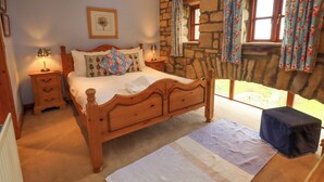 A bedroom with a wooden bed, a nightstand with a lamp, a stone wall, and a window with floral curtains. - Usherwoods Barn (Lancaster)