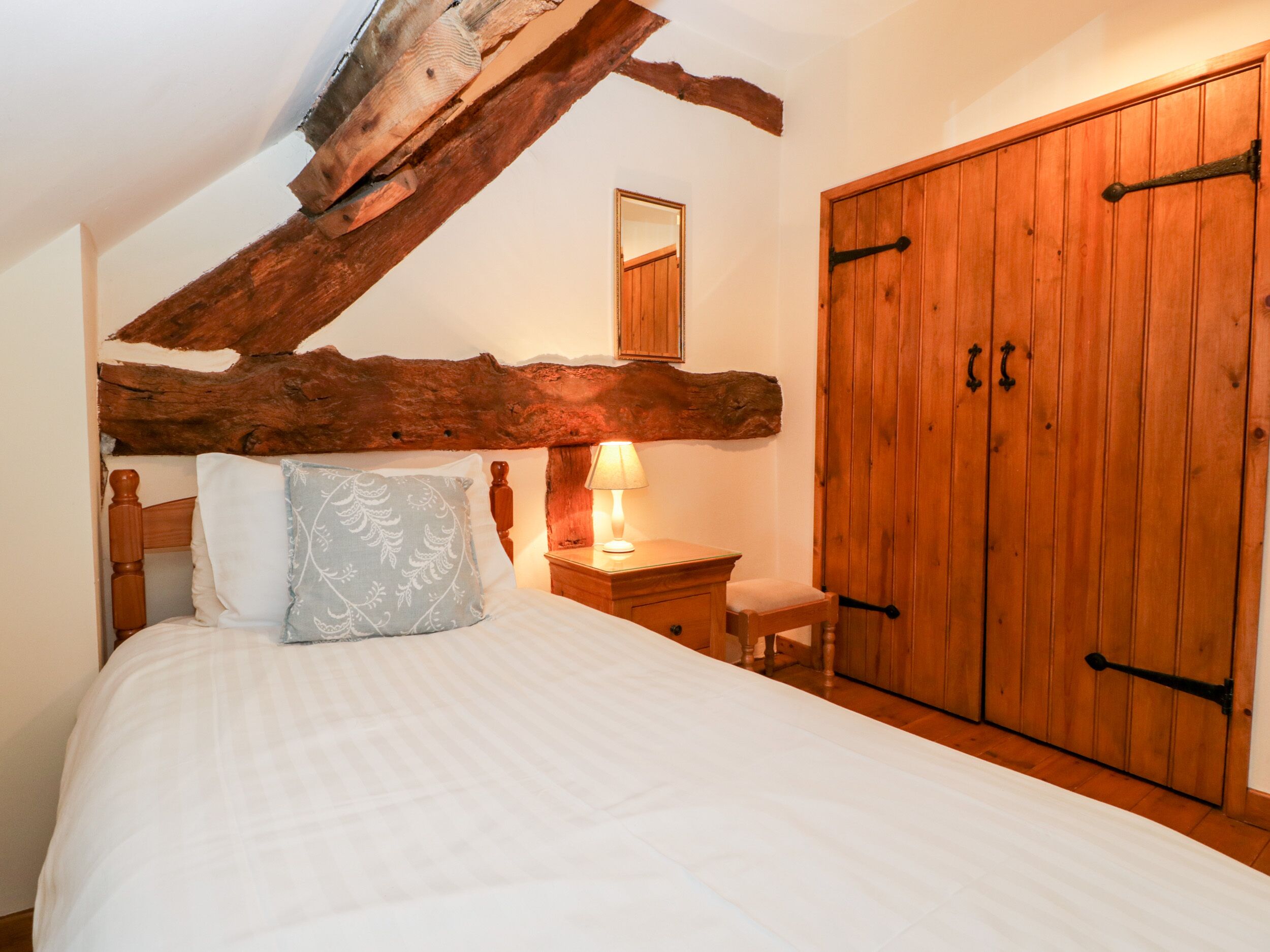 Un dormitorio con viga de madera en el techo, una cama con colcha blanca, una mesita de noche con lámpara y un armario de madera.