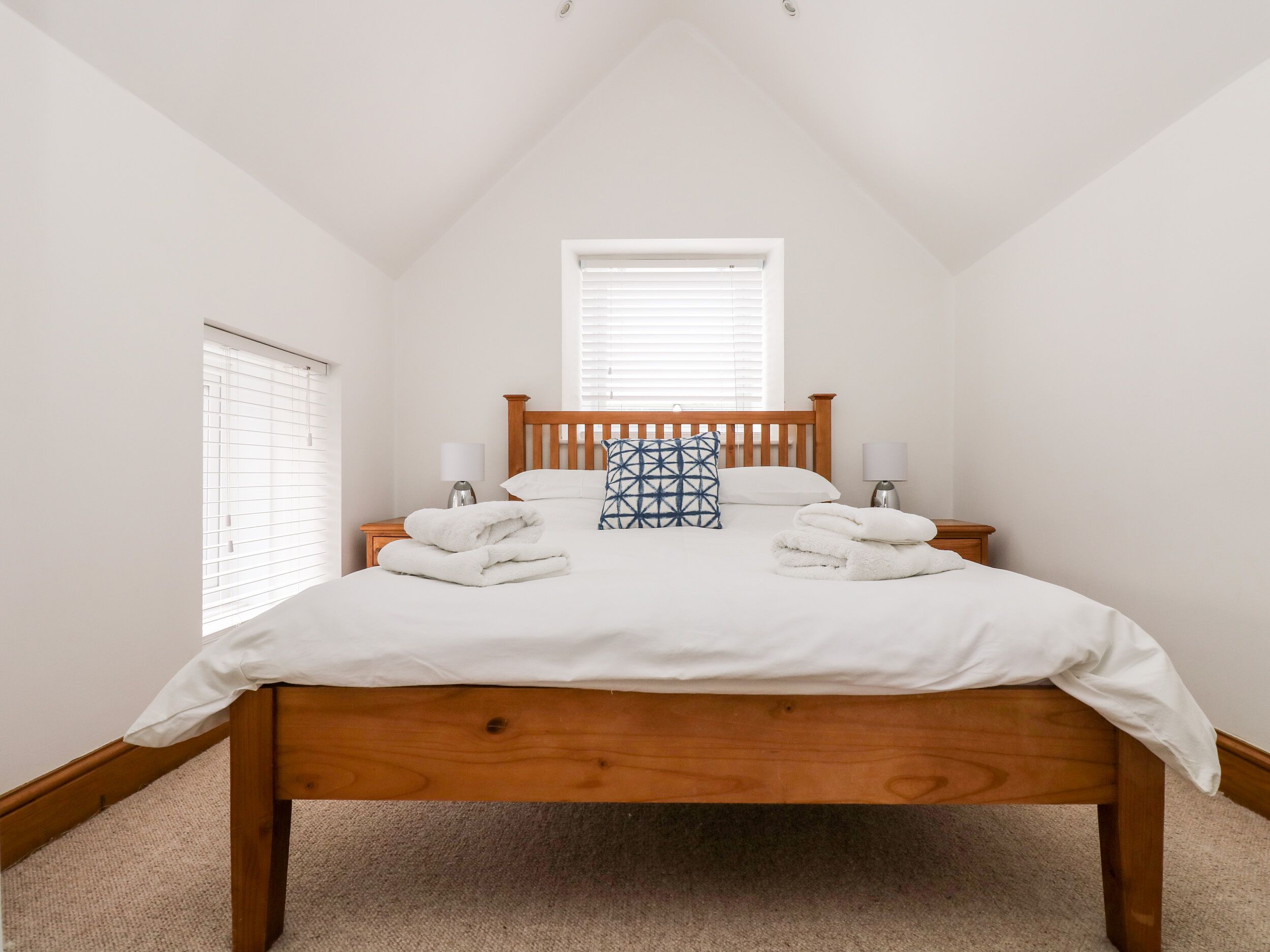 Una cama bien hecha con sábanas blancas y dos almohadas, cabecera de madera y dos lámparas de noche.