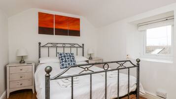 A bedroom with a metal bed frame, white bedding, a nightstand with a lamp, a bedside table, and a window with a view of the roof.