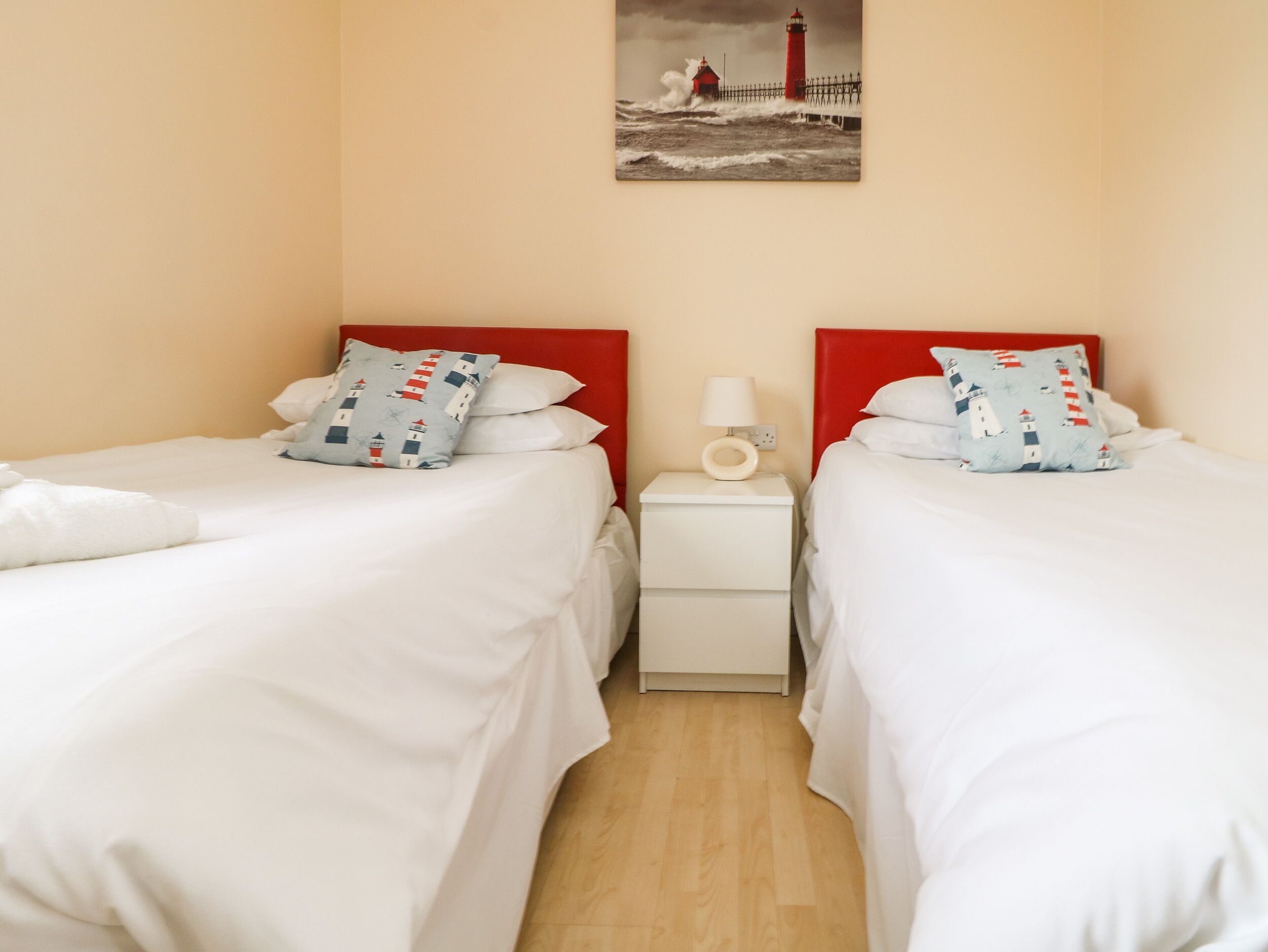 A hotel room with two beds, each with white bedding and red headboards. There is a nightstand with a lamp and a wall-mounted picture of a lighthouse.