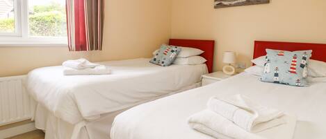 A hotel room with two beds, a window with curtains, a painting of a lighthouse, and a bedside table with a lamp.
