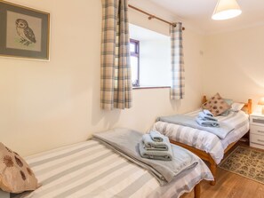 A bedroom with two single beds, a bedside table with a lamp, a window with curtains, and a framed picture on the wall.