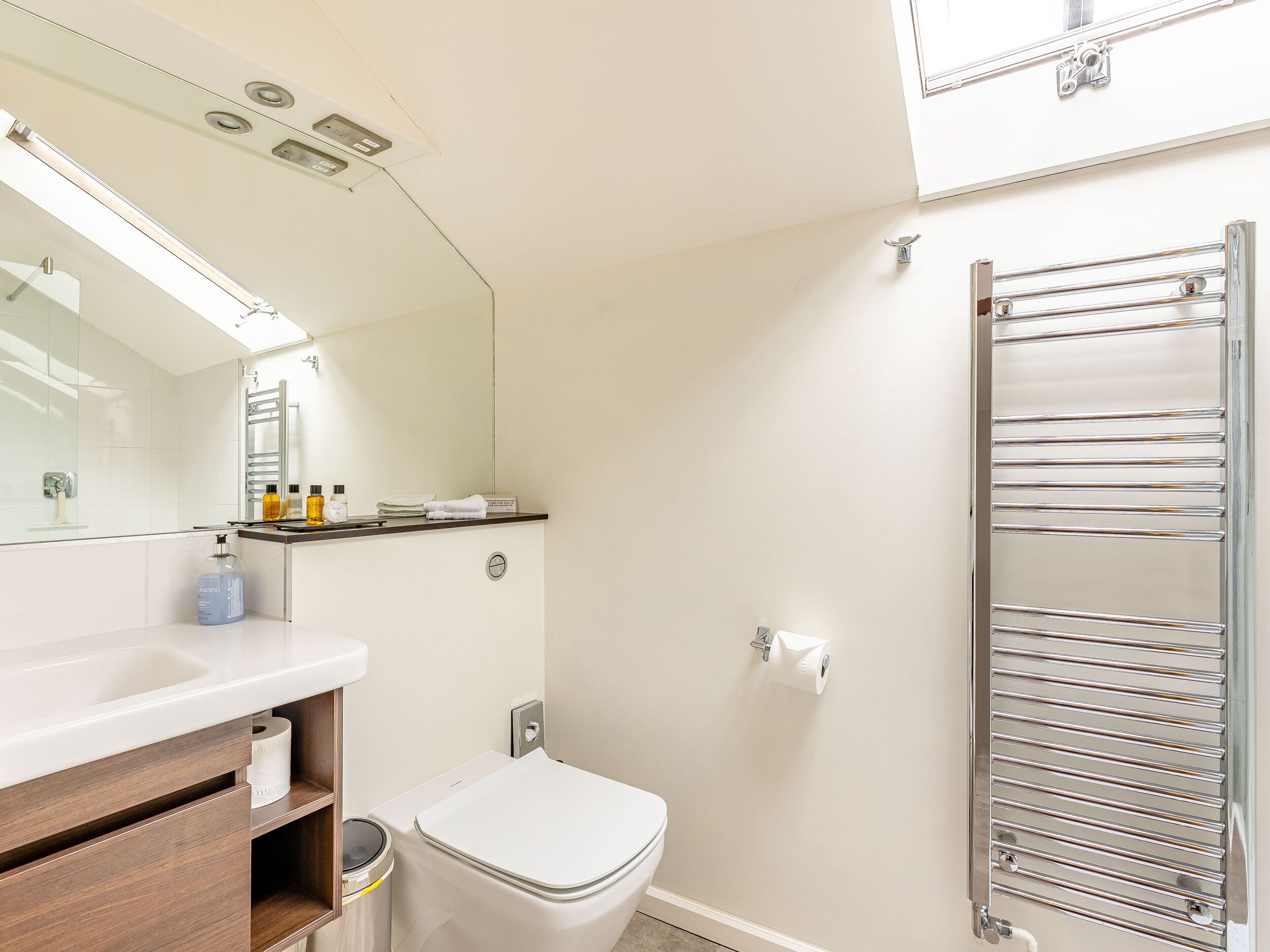 Ein modernes Badezimmer mit einem weißen Waschbecken, Spiegel und Toilette. An der Wand ist ein Handtuchhalter angebracht und auf der Ablage steht eine Flasche Handseife.