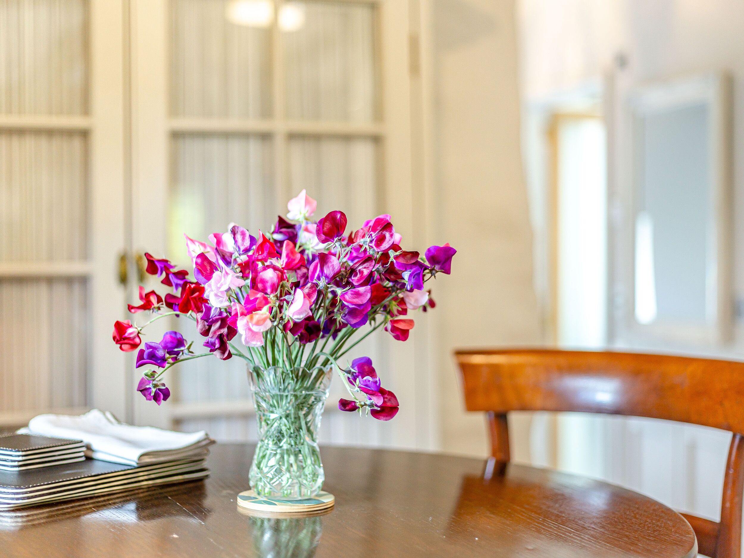 A table with a vase of purple and red flowers, a stack of notebooks, and a wooden chair.