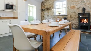 Un comedor con pared de piedra, vigas de madera, chimenea y cocina con armarios blancos y un microondas.