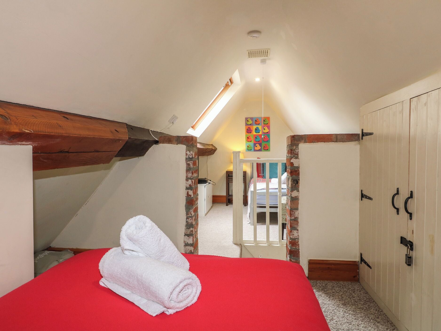 Quarto com colcha vermelha, toalha branca, vigas de madeira, teto inclinado e um ornamento colorido na parede.