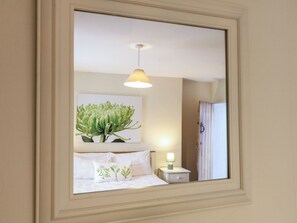 A bedroom with a bed, pillows, a nightstand, a lamp, a wall-mounted painting, and a mirror reflecting the room.