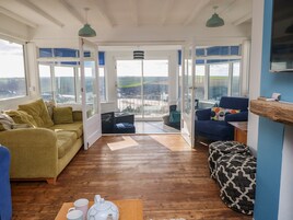 Un salón amplio con piso de madera, un sofá amarillo, un sillón azul y un otomán estampado. Ventanas grandes ofrecen vistas al exterior.