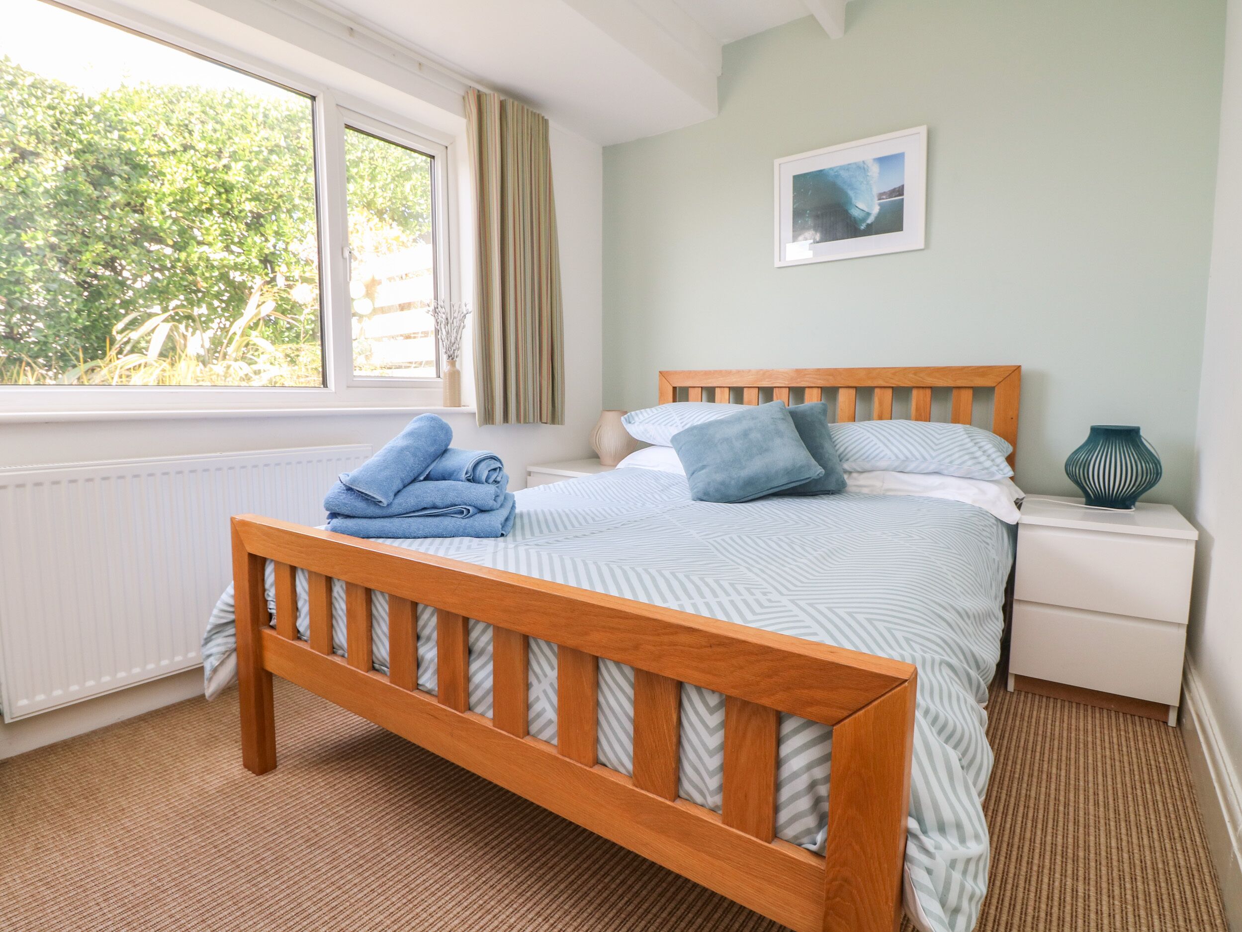 A bedroom with a wooden bed, a nightstand, a window with curtains, and a framed picture on the wall.