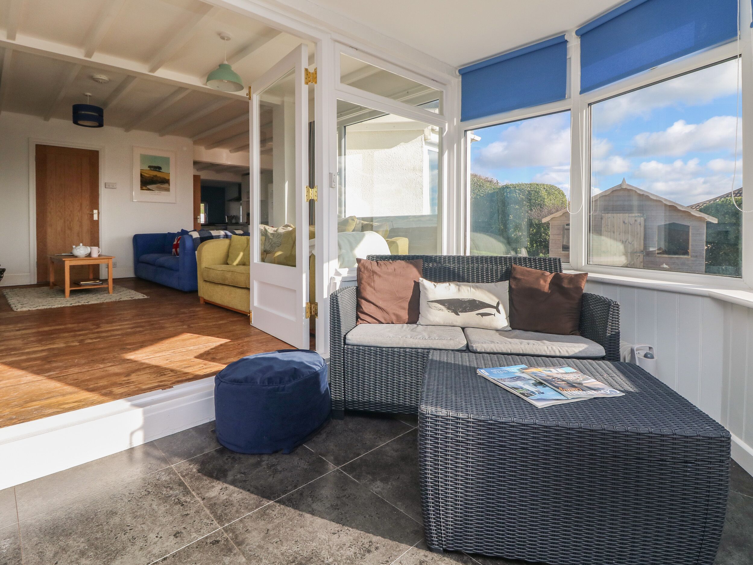 Una sala acogedora con un sofá de mimbre, un reposapiés azul y una mesa de centro de madera. Una puerta da a una habitación luminosa con un sofá amarillo y una mesita.