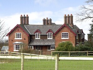 A large brick house with multiple chimneys, a fenced yard, and a small car parked in front. - Dairy Apartment 1 (Knutsford)