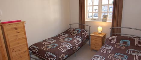A small, well-lit bedroom with two single beds, a wooden dresser, a bedside table with a lamp, and a window with curtains.