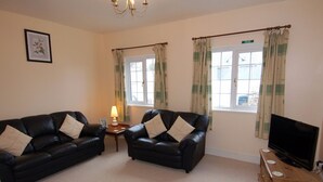 A living room with two black leather sofas, a small table with a lamp, a television on a wooden stand, and two windows with curtains. - The Barn (Camelford)