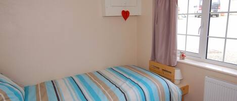 Une petite chambre avec un lit simple, une couverture à rayures, une fenêtre avec des rideaux, un radiateur et un ours en peluche posé sur le rebord de la fenêtre.