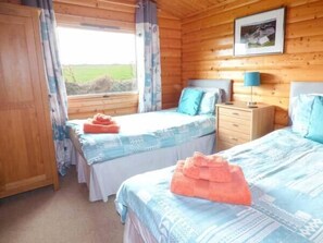 Quarto de cabana de madeira com duas camas, uma mesinha de cabeceira e janela com cortinas.