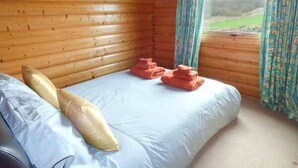A wooden cabin room with a bed, pillows, and a window with curtains. - Ty Pren (Haverfordwest)