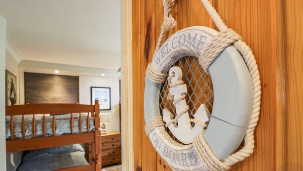 Uma porta de madeira com decoração de corda e âncora, uma cama beliche e uma placa de boas-vindas emoldurada.