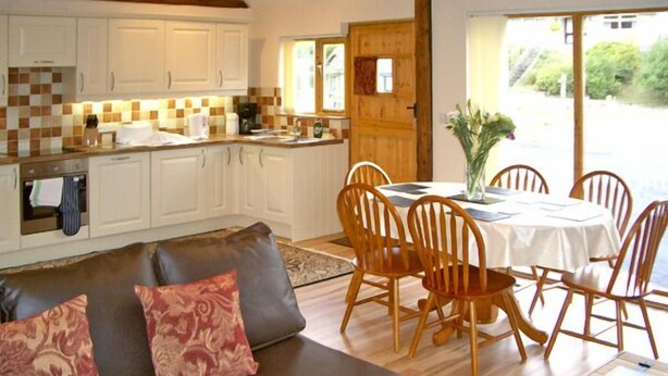 A cozy living room with a brown leather sofa and patterned cushions, a wooden dining table with chairs, and a kitchen with white cabinets and a checkered backsplash. - Happy Union Stables (Llandrindod Wells)