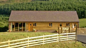 A wooden cabin with a large porch, a fenced yard, and a forested hill in the background. - Happy Union Stables (Llandrindod Wells)