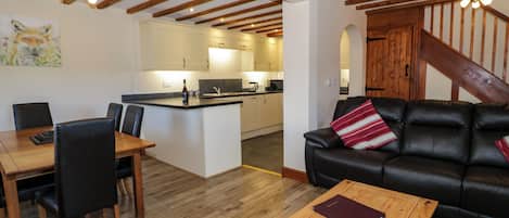 A spacious living area with a wooden dining table, black leather sofa, and a kitchen in the background.