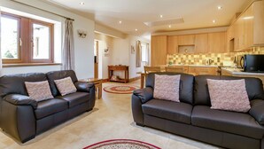 A modern living room with two black leather sofas, a wooden dining table, and a kitchen area with wooden cabinets and a black microwave. - Routster Cottage (Settle)