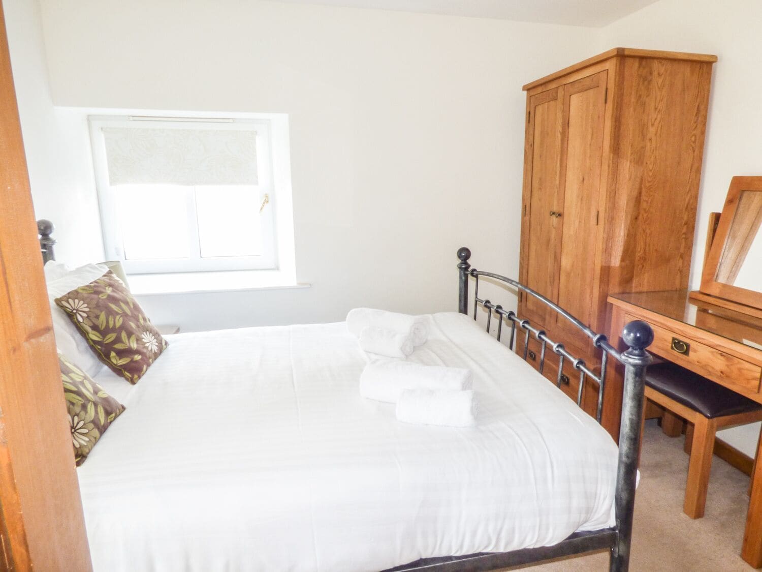 Una cama bien hecha con sábanas y almohadas blancas, un armario de madera y una ventana con persianas.
